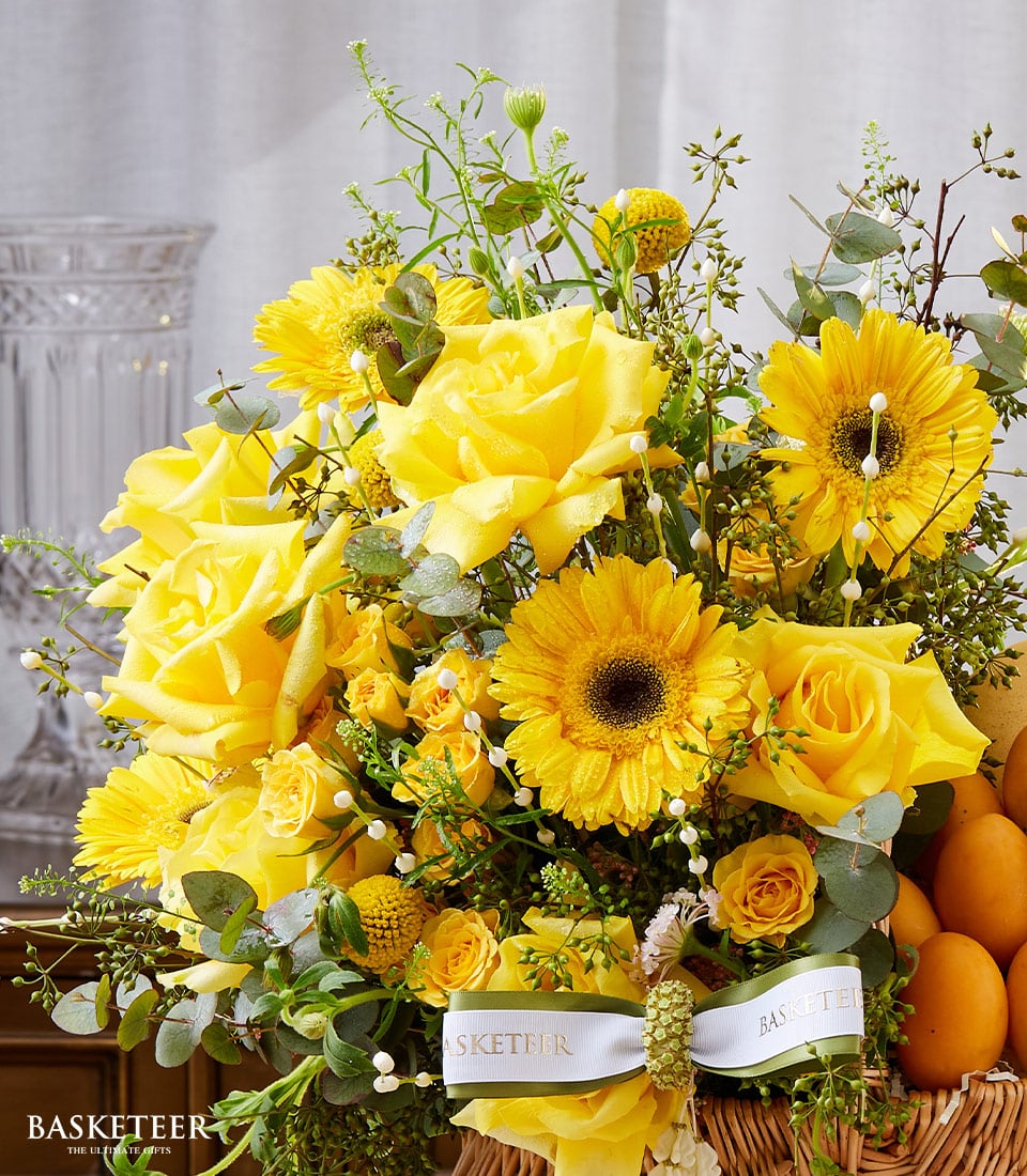 A beautifully arranged, luxurious floral display featuring vibrant yellow roses and cheerful sunflowers, accented with lush mixed greenery, showcased in an elegant wicker basket. The ensemble is complemented by a sophisticated white ribbon elegantly inscribed with the prestigious 