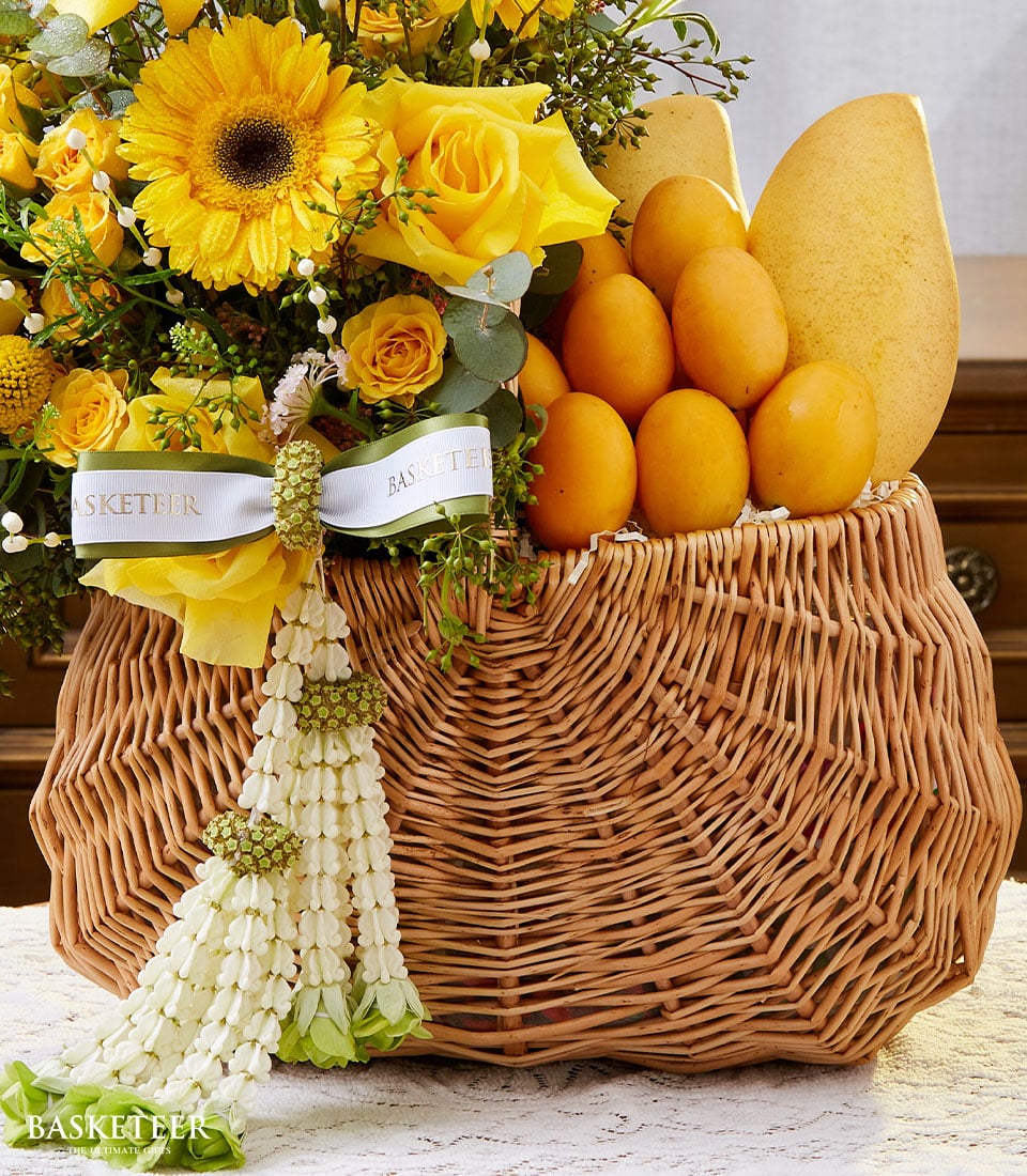 A meticulously crafted wicker basket, elegantly adorned with vibrant yellow flowers, lush green foliage, and pristine white ribbons. Inside this luxurious Basketeer arrangement lie several perfectly ripened golden mangoes, making it an ideal and opulent gift for Songkran festivities. The base of the basket boasts a graceful cascade of delicate white flowers and verdant accents. The distinguished word 