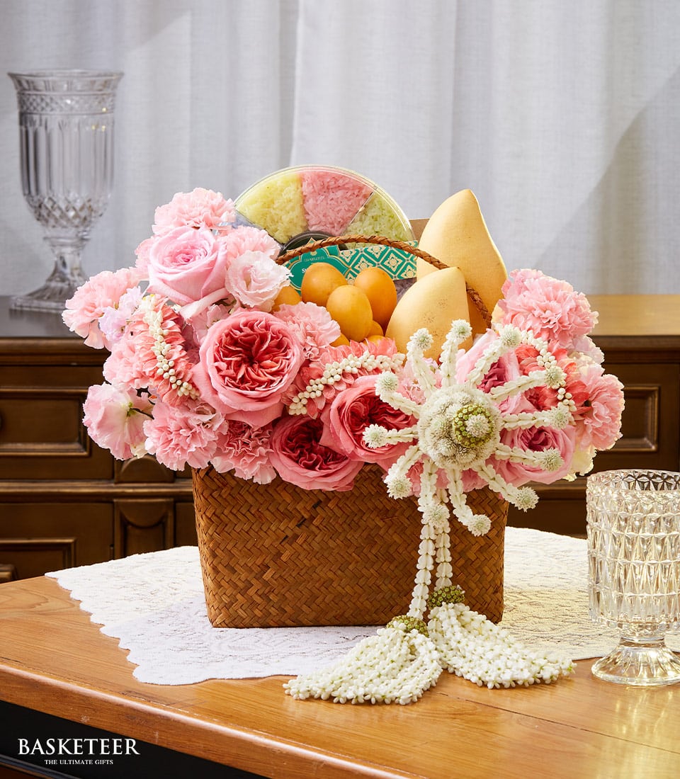 A luxurious woven basket, bearing the distinguished Basketeer logo on an elegantly tied ribbon, cradles a vibrant bouquet of pink roses and carnations. Juicy yellow mangoes and an assortment of colorful jelly sweets nestle amid the blooms. This exquisite arrangement is adorned with a Songkran floral garland and rests gracefully on a polished wooden table, set against the pristine backdrop of white curtains—a sophisticated gift presentation that exudes elegance and charm.