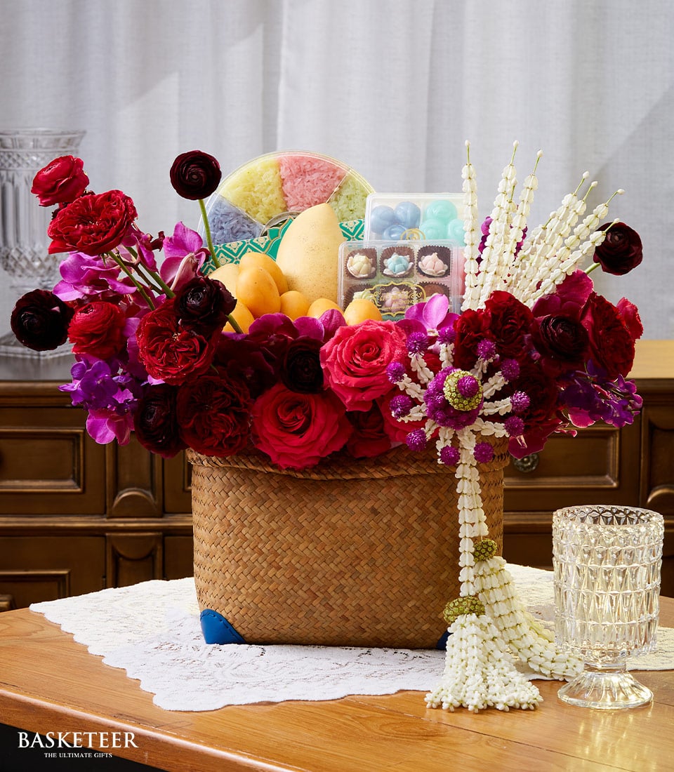 A sumptuous woven basket overflowing with vibrant red, pink, and purple flowers nestles alongside luscious mangoes and an array of colorful traditional sweets. This exquisite arrangement sits atop a lace-covered wooden table, accented with an elegant floral garland and a sparkling crystal glass. The distinguished Basketeer brand logo graces the scene, epitomizing the perfect luxurious gift for Songkran.