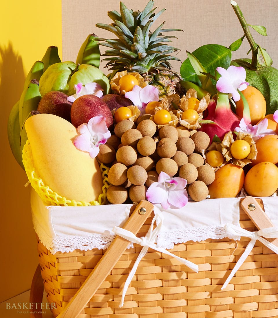 A stunning wicker basket, exquisitely assembled by Basketeer, overflowing with a vibrant assortment of tropical fruits including a perfectly ripe pineapple, succulent bananas, juicy mangoes, delicate lychees, luscious peaches, and tantalizing passion fruits. This luxurious Thai Summer Orchard Basket is elegantly adorned with refined white lace and complemented by an artful arrangement of delicate pink and white flowers. The scene is set against an inviting backdrop of warm yellow and beige tones that enhance the opulence of this delightful ensemble.