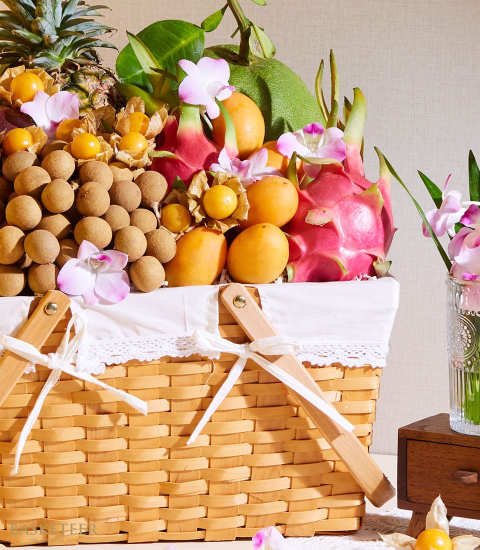 The Basketeer Thai Summer Orchard Basket, an exquisite collection of tropical fruits including dragon fruits, longans, loquats, and a perfectly ripe pineapple. This luxurious wicker basket features a pristine white fabric edge and is artfully adorned with delicate pink and white flowers. The elegant arrangement sits invitingly on a light background, exuding sophistication and opulence. Basketeer branding is subtly integrated into the ribbon tying the ensemble together, ensuring an unforgettable touch of class.