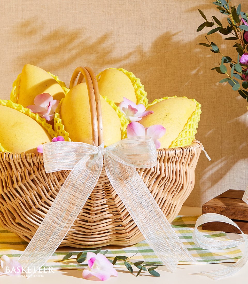 A luxurious wicker basket brimming with succulent Organic Golden Mangoes, each cradled in elegant yellow protective netting. The basket is beautifully adorned with a pristine white ribbon inscribed with the Basketeer logo, and is surrounded by an artful arrangement of pink and white blossoms. In the background, a roll of matching white ribbon and lush green leaves add to the opulent ambiance, inviting you to indulge in nature's finest offerings from Basketeer.