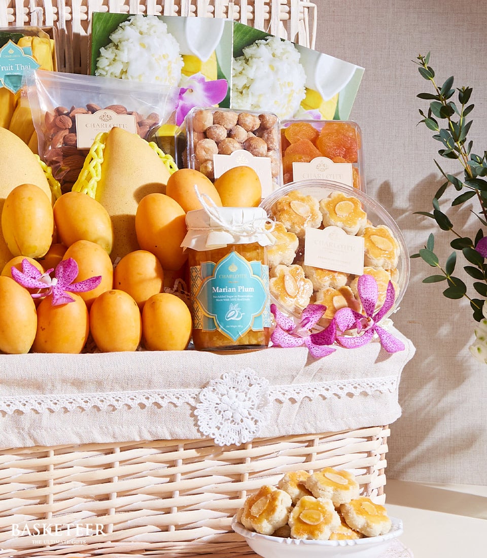 A sumptuous cream-colored wicker basket, brimming with an exquisite selection of golden fruits and gourmet packaged snacks. Featured items include succulent mangoes, radiant golden berries, premium nuts, delectable candied fruits, and a jar of luxurious Marian plum jam. The presentation is graced with delicate decorative flowers that add a touch of elegance to the Thai Dessert-Fruits Gift Basket by Basketeer. In front of the basket sits a petite dish showcasing an array of these delightful treats. A ribbon bearing the Basketeer logo ties everything together in luxurious harmony, making this gift both opulent and inviting.