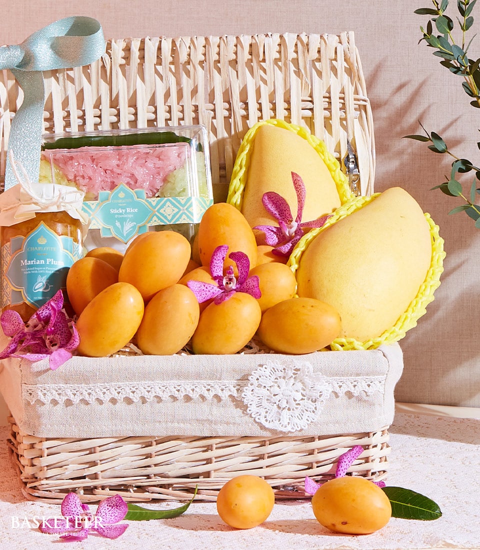 A lavishly designed wicker basket brimming with ripe Thai organic mangoes, fragrant sticky rice, dried flowers, and a jar of artisanal jam graces a table adorned with delicate lace. The tropical-themed packaging is elegantly complemented by vibrant purple orchid flowers. Atop the basket lid, a sumptuous bow showcases the elevated presentation of Basketeer.