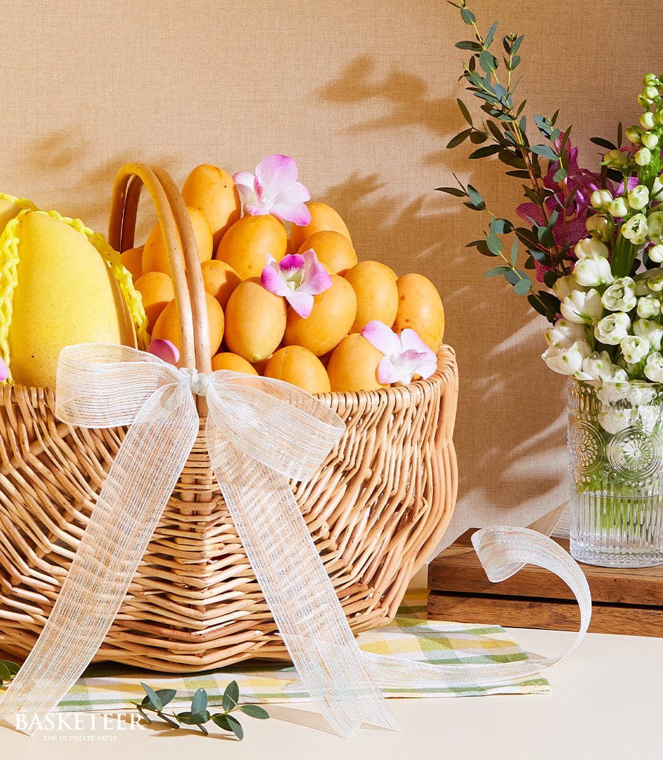 A luxurious wicker basket, elegantly branded with a ribbon featuring the Basketeer logo, overflows with an assortment of premium organic mangoes and Marian plums. The bounty is beautifully adorned with delicate pink flowers and a pristine white ribbon, all nestled atop an inviting plaid cloth. Beside the basket, a crystal-clear vase showcases a striking bouquet of vibrant purple and white flowers, creating an enchanting scene against a sophisticated beige backdrop.