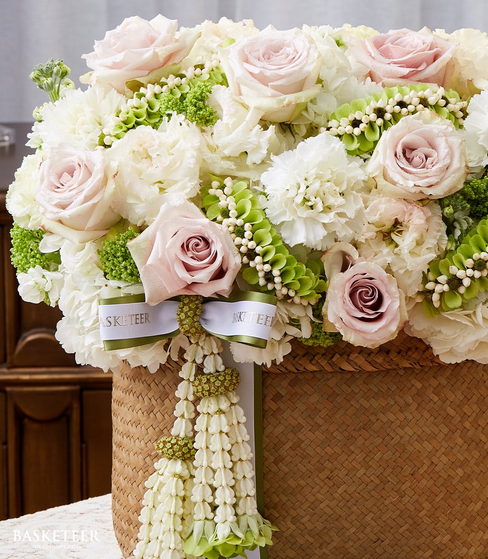 A luxurious woven basket brimming with an exquisite arrangement of pale pink roses, pristine white peonies, lush green hydrangeas, and other delicate Thai classic flowers. Elegant white ribbons bearing the 
