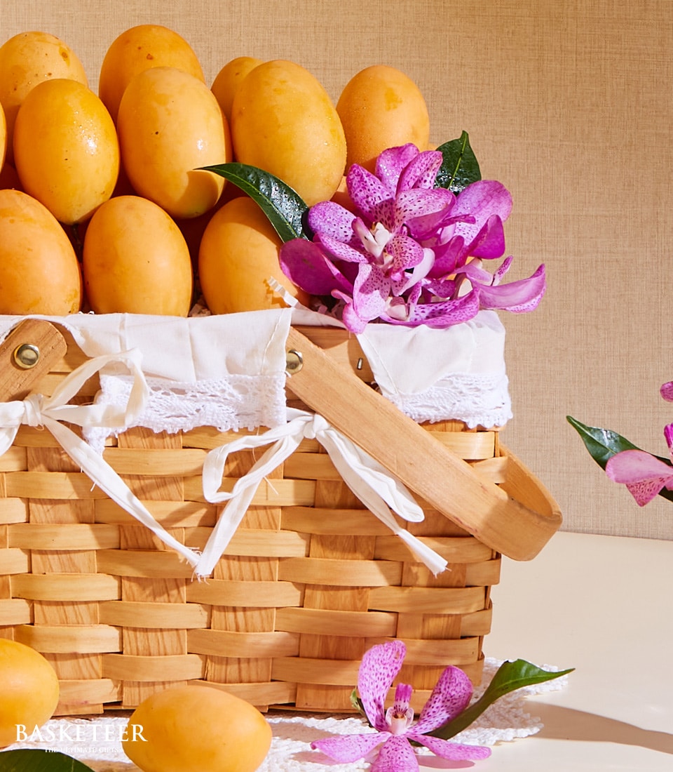A lavish wicker basket from Basketeer brimming with succulent yellow mangoes and exotic marian plum fruits, elegantly lined with a delicate white fabric featuring intricate lace detailing. Striking purple orchids are impeccably arranged around the basket, enhancing its opulent appearance. The wooden handle is gracefully tied with a luxurious ribbon adorned with the Basketeer logo. Set against a sophisticated neutral beige backdrop, this scene exudes luxury and invites indulgence.