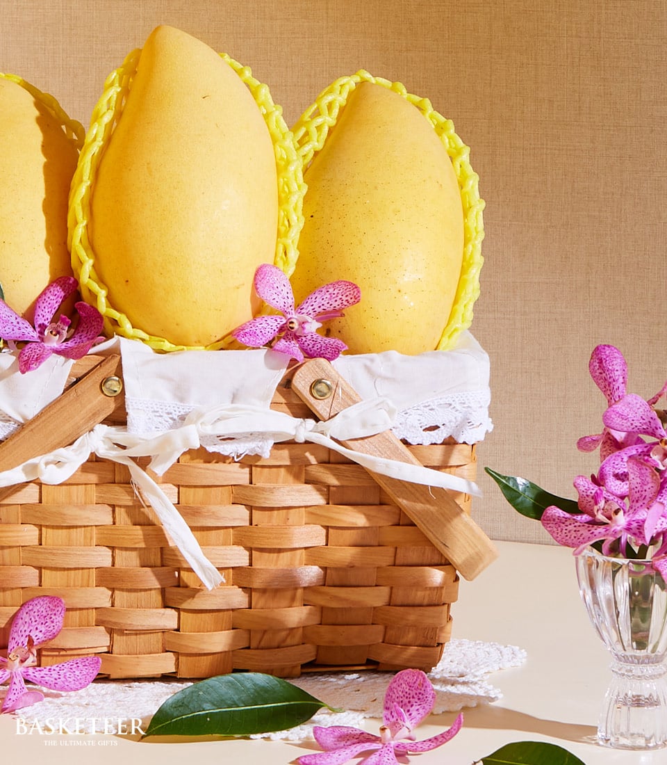 A luxurious wicker basket overflowing with three sumptuous yellow Marian Plum fruits, each meticulously wrapped in an elegant yellow protective sleeve. The basket is elegantly lined with a pristine white cloth and exquisitely adorned with delicate pink orchid blossoms. Additional vibrant pink orchids are artfully arranged around the basket and showcased in a nearby petite glass vase, capturing the essence of opulence. Look for the distinguished Basketeer logo subtly embossed on a ribbon, symbolizing unparalleled quality and sophistication.