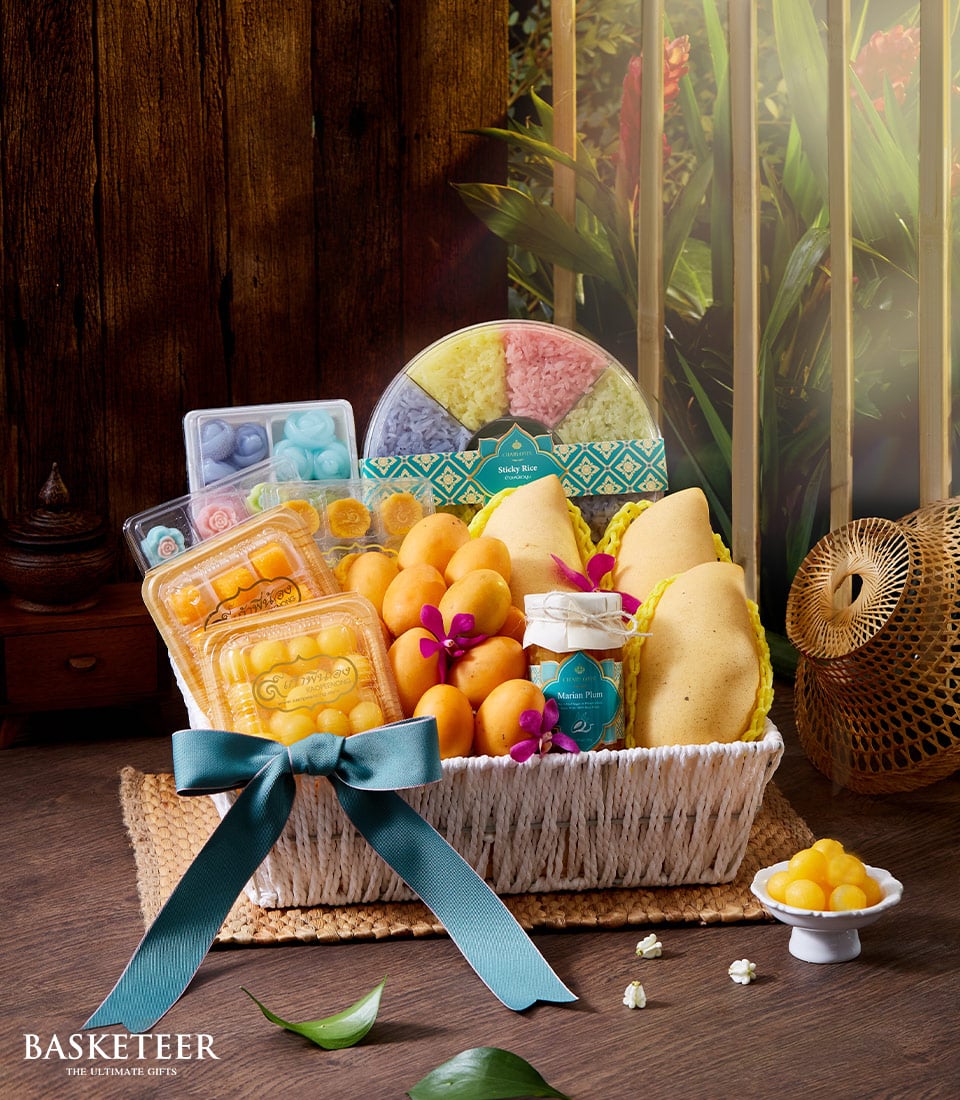 A beautiful wicker gift basket adorned with a luxurious teal bow from Basketeer showcases an exquisite assortment of premium fruits, vibrant sweets, and an authentic Thai tropical dessert. Elegantly arranged on a polished wooden table, the scene is further enhanced by the warm glow of a lantern and the serene greenery visible through the window. This inviting setting radiates charm, making it an irresistible offering from Basketeer.