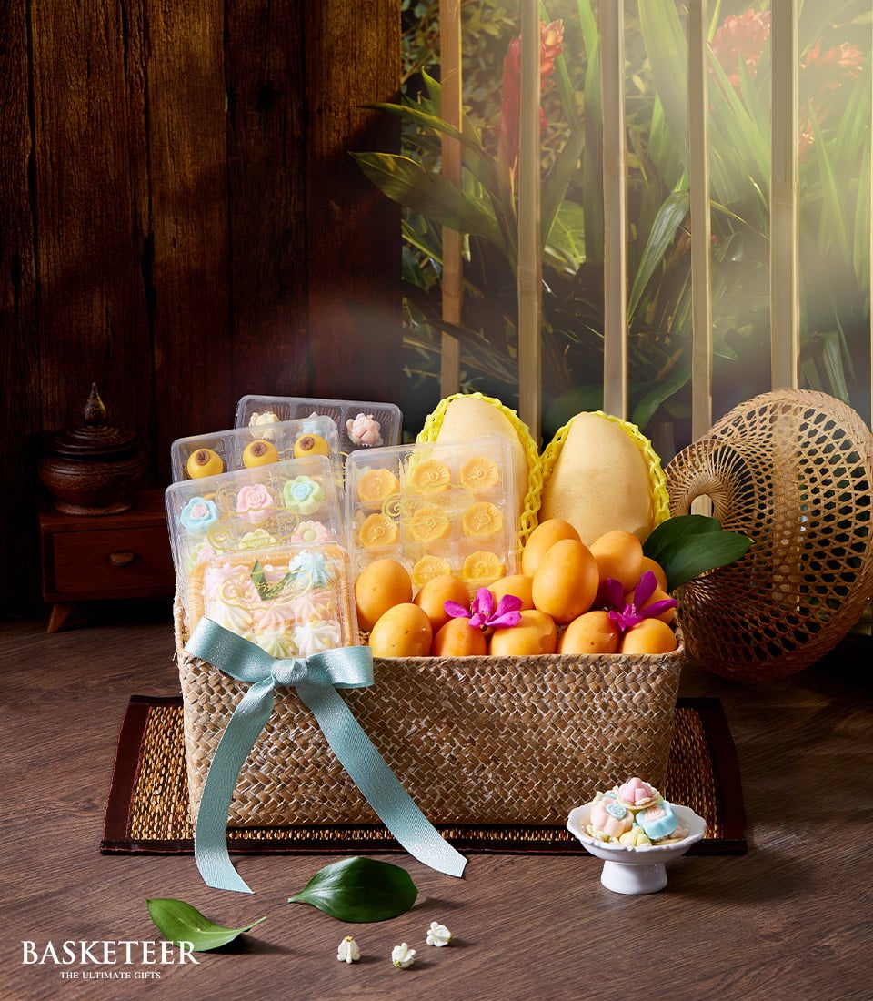 A luxurious Yellow Fruits & Thai Dessert Basket by Basketeer, overflowing with succulent fresh yellow mangoes and elegantly adorned with intricately crafted sweets, sits magnificently on a polished wooden surface. An exquisite light blue ribbon featuring the Basketeer logo cascades around the basket, accentuating its opulence. The lush greenery visible through the wide window creates an enchanting backdrop, while a charming wicker hat and an artfully arranged flower-shaped dessert add a touch of sophistication to this inviting scene.
