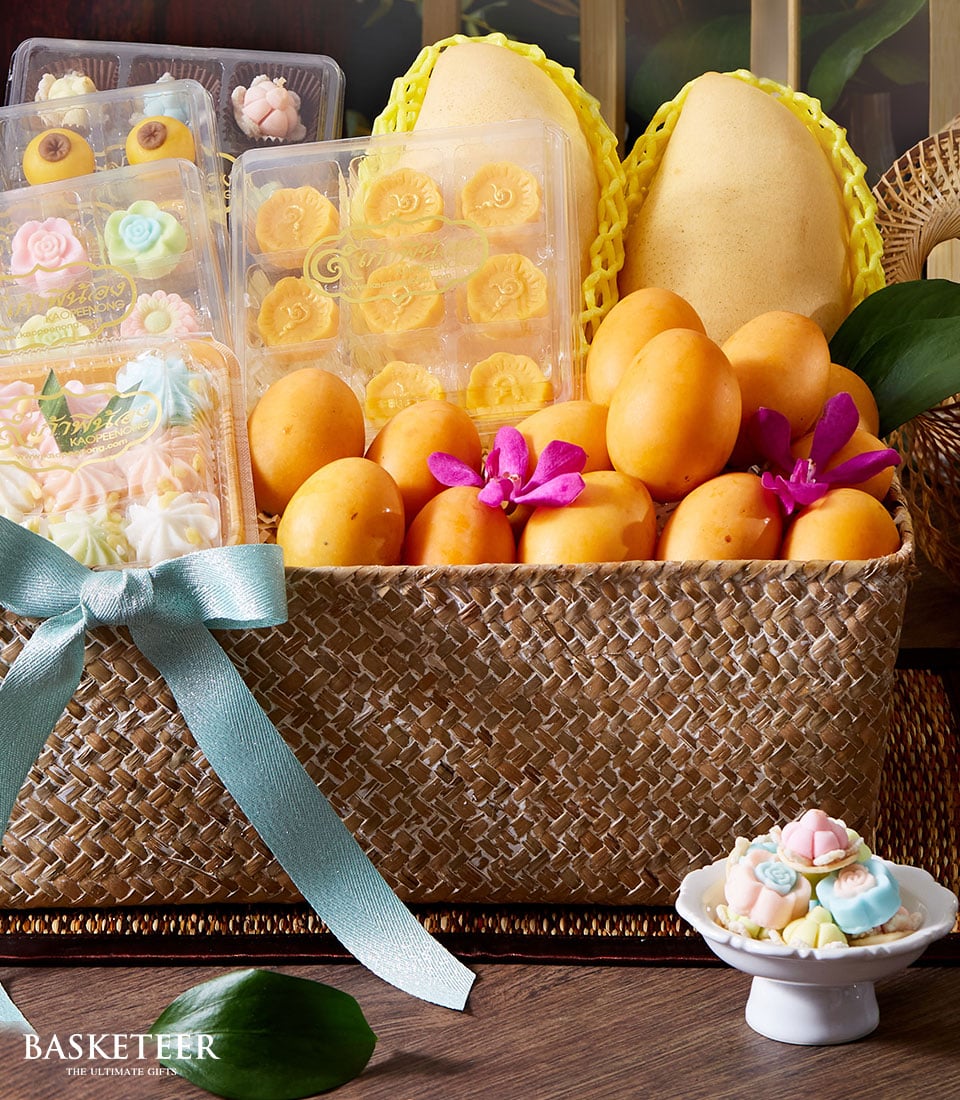 A luxurious wicker basket brimming with vibrant, golden mangoes and exquisitely arranged flower-shaped Thai pastries awaits your indulgence. Additional delicately netted mangoes are nestled towards the back of the basket. The opulent Thai Dessert Basket is elegantly adorned with a light blue bow and proudly features the Basketeer logo on a tastefully positioned ribbon. In front of this sumptuous ensemble, an elegant dish presents a colorful array of intricate floral desserts, inviting you to savor each delightful bite in true Basketeer fashion.