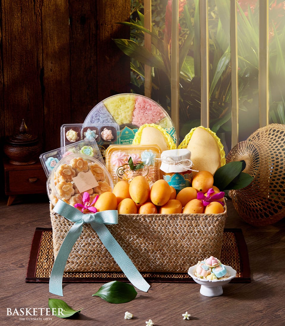 A luxurious woven basket, overflowing with an exquisite selection of tropical fruits such as ripe mangos, accompanied by an array of delightful treats and delectable Thai desserts. The Thai Fruits and Dessert Gift Basket from Basketeer is elegantly adorned with a light blue ribbon, surrounded by enchanting decorative elements including vibrant orchids and charming small porcelain pieces. The prestigious Basketeer logo graces the scene, adding a touch of sophistication to this opulent gift offering.