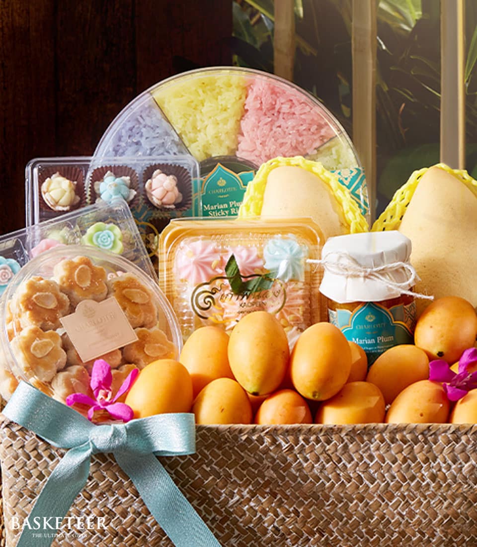 A lavish wicker basket overflowing with succulent, golden-yellow Marian plums takes center stage, elegantly adorned with a delicate light blue ribbon. Encompassed by the fresh bounty are an array of gourmet delights: meticulously packaged dried fruits, vibrant Thai desserts, a jar of artisanal jam, and exquisite decorative flowers. A charming box proudly displays the 