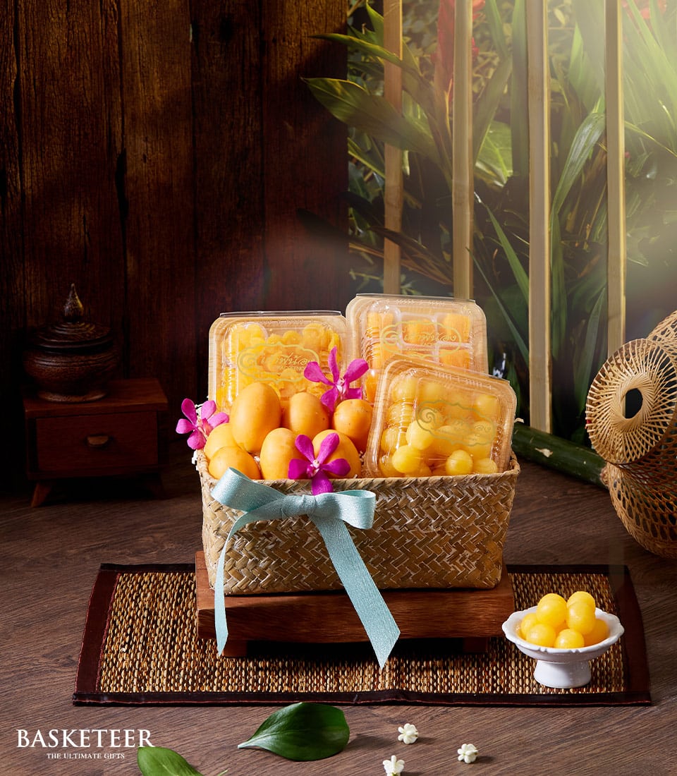 A luxurious Basketeer basket brimming with an exquisite selection of yellow fruits and decadent Thai sweets is elegantly perched on a polished wooden pedestal. The basket, adorned with a sophisticated blue ribbon and vibrant purple flowers, radiates opulence. Nearby, a delicately crafted bowl overflows with additional yellow sweets, inviting indulgence. This tropical tableau captures the essence of Songkran in the most enchanting manner. Adorned with the Basketeer logo, this setting exudes exclusivity and invites you to savor every delightful moment.