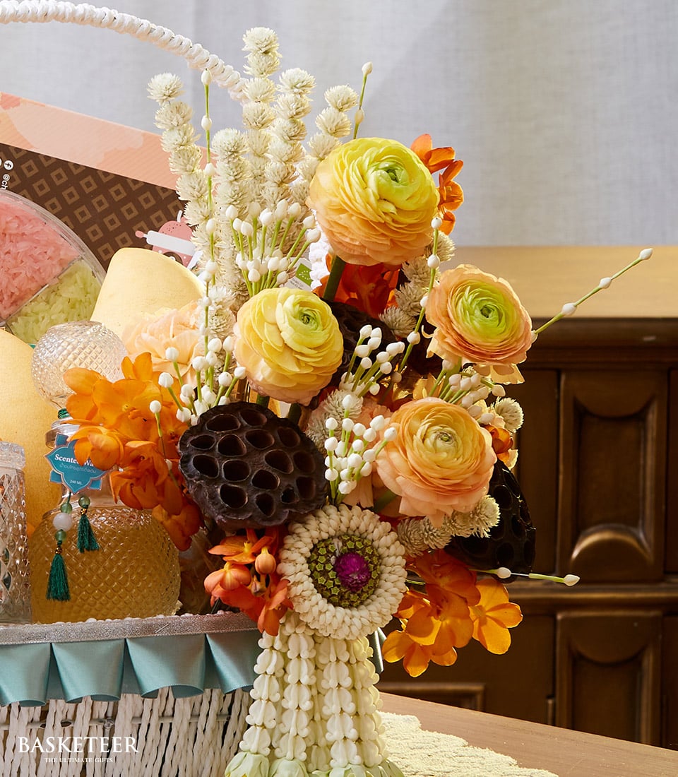 An exquisite display featuring vibrant yellow and orange ranunculus flowers, elegantly paired with dried pods and ornamental accents, all beautifully arranged in a woven white basket. The Thai Flowers & Dessert Gift Basket by Basketeer exudes luxury and charm as part of an opulent gift set, with tantalizing delights peeking invitingly from the background. A satin ribbon adorned with the Basketeer logo graces the arrangement, adding an extra touch of sophistication to this indulgent offering.