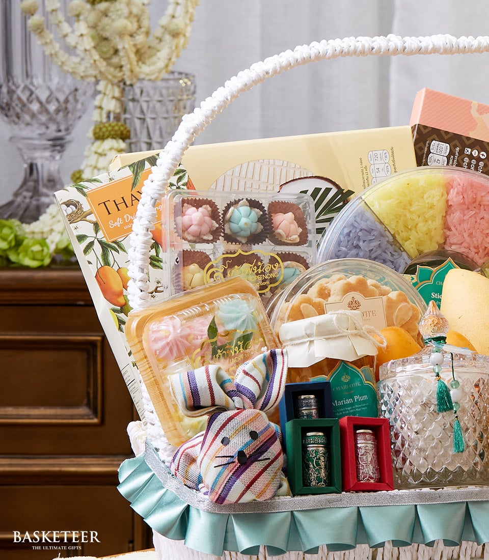 A lavish close-up of the Basketeer Thai Flowers & Dessert Gift Basket, a treasure trove of opulent delights. This exquisite basket overflows with vibrant bath bombs, an adorable striped fabric bunny, elegant tea canisters, a chic box of assorted sweets bearing the Basketeer insignia, and other indulgent luxuries. Adorned with a sumptuous green satin ribbon emblazoned with the Basketeer logo, this enticing arrangement sits invitingly on a polished wooden surface.
