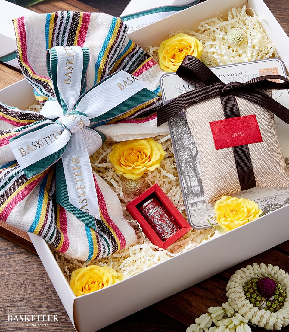 A luxurious gift box from Basketeer showcases an array of colorful striped fabrics, elegantly paired with fresh, vibrant yellow roses. Nestled beside them is a small red container holding a gleaming metal object, enhancing the opulent display. The scene is further elevated by the aromatic delight of the Songkran Incense Gift Set. Completing this exquisite arrangement is a tan pouch adorned with a sophisticated red label and tied gracefully with a brown ribbon, all set against shredded paper bearing the distinguished 