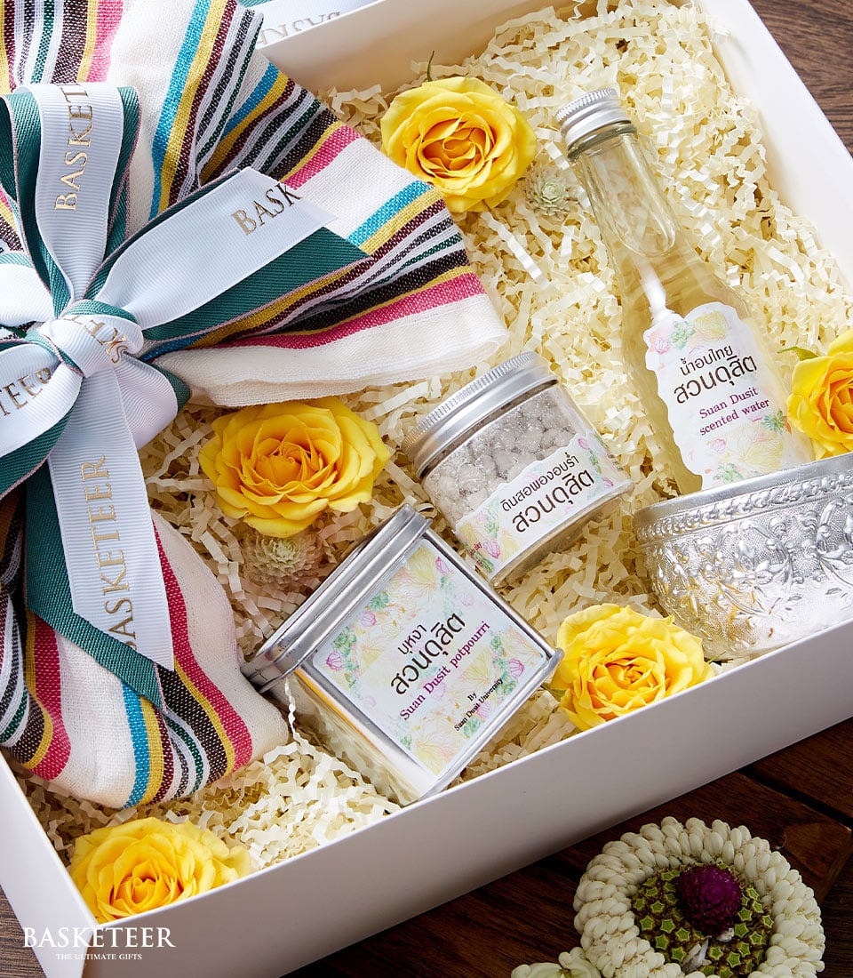 A Basketeer Thai Traditional Perfume Gift Set exudes opulence and charm, wrapped in a colorful striped bow. The presentation is adorned with vibrant yellow roses, adding a touch of elegance. Inside the luxurious box filled with finely shredded paper rests an exquisite collection: a delicate bottle of fragrant liquid, a beautifully crafted tin container, a petite tin for added allure, and an intricately designed silver bowl. Outside the box on the rich wooden surface lies an ornate flower decoration, completing this lavish ensemble by Basketeer.