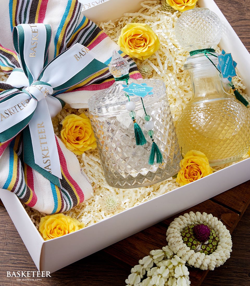 A lavish Basketeer gift box unveils a stunning display: a bottle and elegantly patterned glass container filled with exquisite yellow Thai traditional perfume. Nestled amidst delicate shredded paper and adorned with radiant yellow roses, this ensemble exudes opulence. A chic striped cloth, meticulously tied with a ribbon inscribed with 