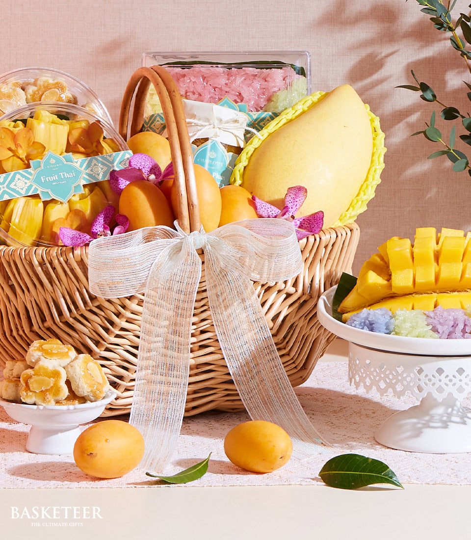 A luxurious wicker basket, elegantly adorned with a pristine white ribbon showcasing the Basketeer logo, brims with an exquisite assortment of fresh fruits and golden-hued snacks. Nestled within is a transparent box of vibrant Thai desserts that add a splash of color to the ensemble. Juicy mangoes are artfully displayed beside this sumptuous summer fruit collection. Nearby, a sophisticated cake stand presents intricately prepared sticky rice in captivating hues, epitomizing culinary artistry and inviting indulgence.