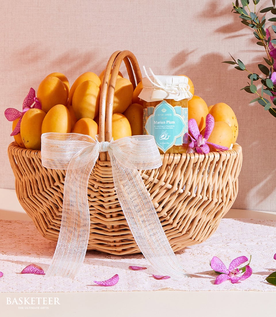 The Sweet Marian Plum Basket by Basketeer exudes luxury and elegance with its artisanal wicker basket brimming with luscious, golden marian plums, complemented by an exquisite jar of marian plum preserve tied with a pristine white ribbon showcasing the Basketeer logo. Enchanting pink orchids are thoughtfully scattered around the arrangement, their vibrant hue contrasting beautifully against the sophisticated beige background in this radiant and inviting display.