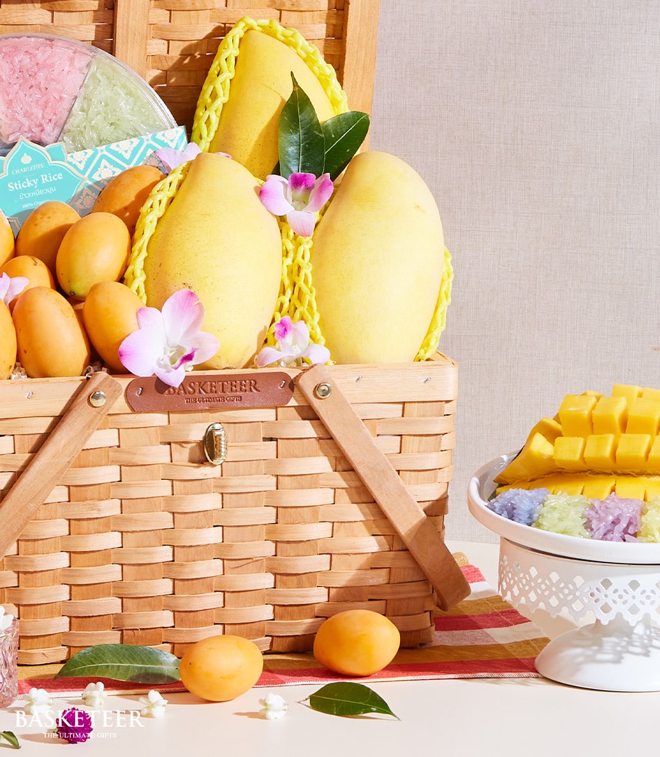 A lavish wicker basket from Basketeer is filled with sun-kissed Thai mangoes, adorned with exquisitely wrapped sticky rice packages in a vibrant array of colors. The arrangement is complemented by delicate flowers, all beautifully presented on an elegant checkered cloth. Nearby, a pristine white pedestal dish showcases perfectly sliced mangoes and an enticing selection of multicolored sticky rice. A subtle ribbon featuring the Basketeer logo adds a touch of sophistication to the inviting scene.