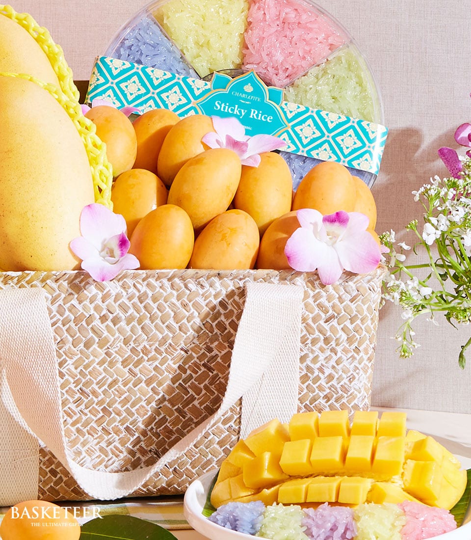 A sumptuous woven basket overflowing with luscious ripe mangoes and an exquisitely packaged assortment of colorful sticky rice, elegantly presented by Basketeer. Next to this delightful arrangement, a perfectly sliced mango and a delicate sprig of pink flowers rest invitingly. The scene is set against a backdrop of soft, light-colored fabric and an opulent bouquet of white and pink blossoms. The word 