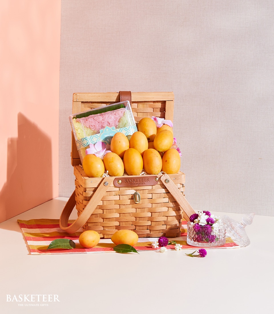 A luxurious wicker basket brimming with golden, succulent mangoes rests atop an elegant striped cloth. This exquisite Marian Plum & Sticky Rice Basket is enhanced by a charming gift wrapped in vibrant green floral paper. Surrounding the basket are additional ripe mangoes and delicate white and purple flowers, thoughtfully arranged in a decorative glass holder. The distinguished 