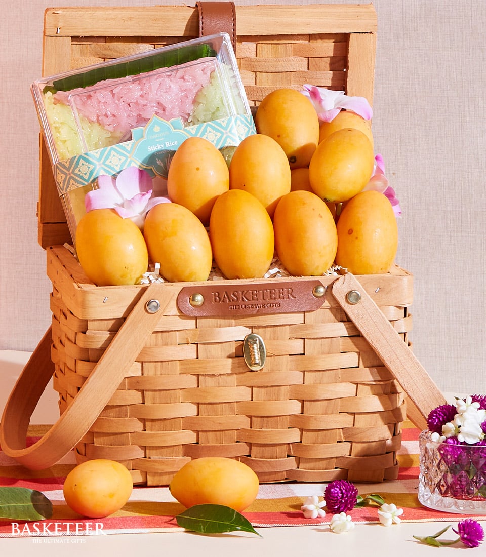 A luxurious woven Marian Plum & Sticky Rice basket, overflowing with perfectly ripe and golden Basketeer mangoes, is elegantly presented. Nestled among the succulent fruits is a premium packet of sticky rice. Delicate pink and white flowers add an exquisite touch to the arrangement. Nearby, a charming floral bouquet and artfully sliced mango rest on a vibrant cloth, enhancing the inviting display. The Basketeer logo subtly adorns a ribbon tied around the basket, symbolizing quality and elegance.
