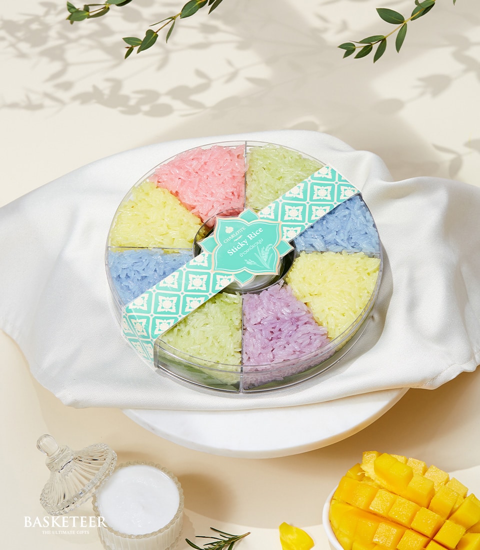 A luxurious Charlotte Moon Sticky Rice Gift by Basketeer, elegantly presented in a round, clear container delicately partitioned into eight sections. Each segment boasts vibrantly hued sticky rice in shades of pink, green, yellow, blue, purple, and white. This indulgent assortment rests on an immaculate white cloth. Nearby, a pristine white cup and refined glass container enhance the opulence of the scene. Delectably sliced mangoes are artfully placed to complete this lavish arrangement. Look closely to discover subtle touches of the Basketeer brand—perhaps on a graceful ribbon or box nearby—reaffirming its commitment to exquisite quality and attention to detail.