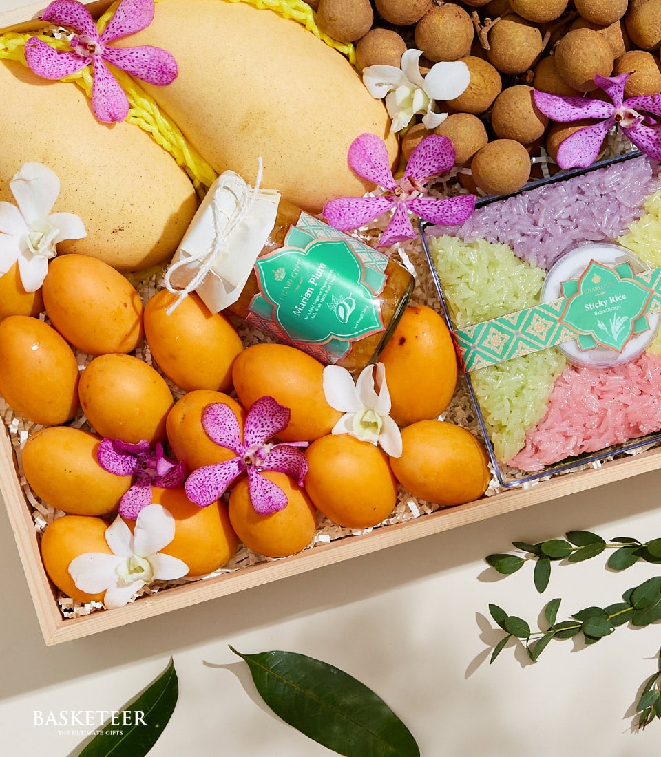 A luxurious wooden tray brimming with freshly-picked mangoes, exotic dragon fruit, succulent longan, and a vibrant container of sticky rice adorned with delicate purple and white blossoms. Each meticulously arranged item within the Basketeer 