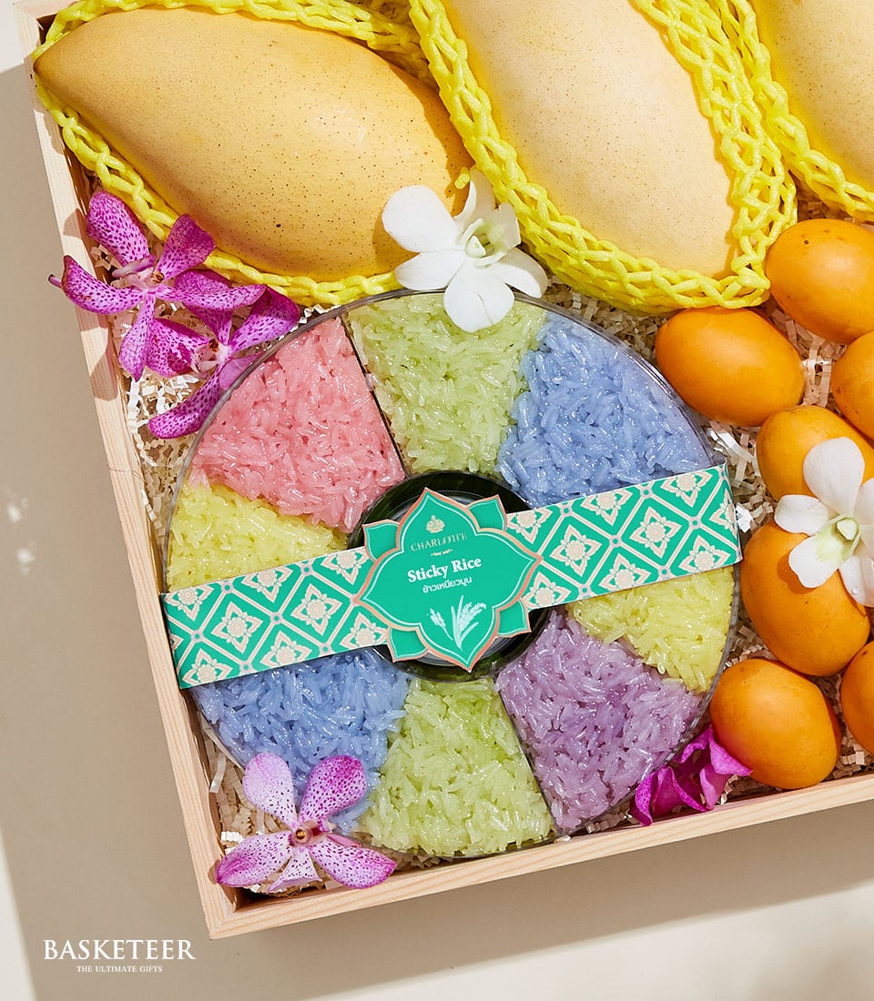 A luxurious wooden gift box from Basketeer showcases an exquisite assortment that invites indulgence. At the pinnacle, three sun-kissed yellow mangoes promise succulent sweetness. The centerpiece features a beautifully circular arrangement of vibrant sticky rice in six enchanting hues, each segment adding a splash of color and rich flavor. Nestled strategically around this masterpiece are several petite Marian Plums, their bright orange skins contrasted by delicate pink and white blossoms. The Basketeer logo elegantly graces the label on the sticky rice, affirming the brand’s dedication to opulence and quality.