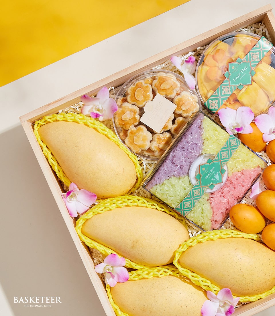 Discover the Thai Summer Harmony Box by Basketeer, an exquisite wooden gift box brimming with succulent ripe yellow mangoes, a medley of vibrant traditional sweets, and delicate small yellow fruits. Meticulously adorned with elegant pink orchids for a touch of sophistication. This luxurious offering is elegantly presented on a radiant yellow surface, proudly showcasing the Basketeer logo in the bottom left corner to signify its brand excellence.