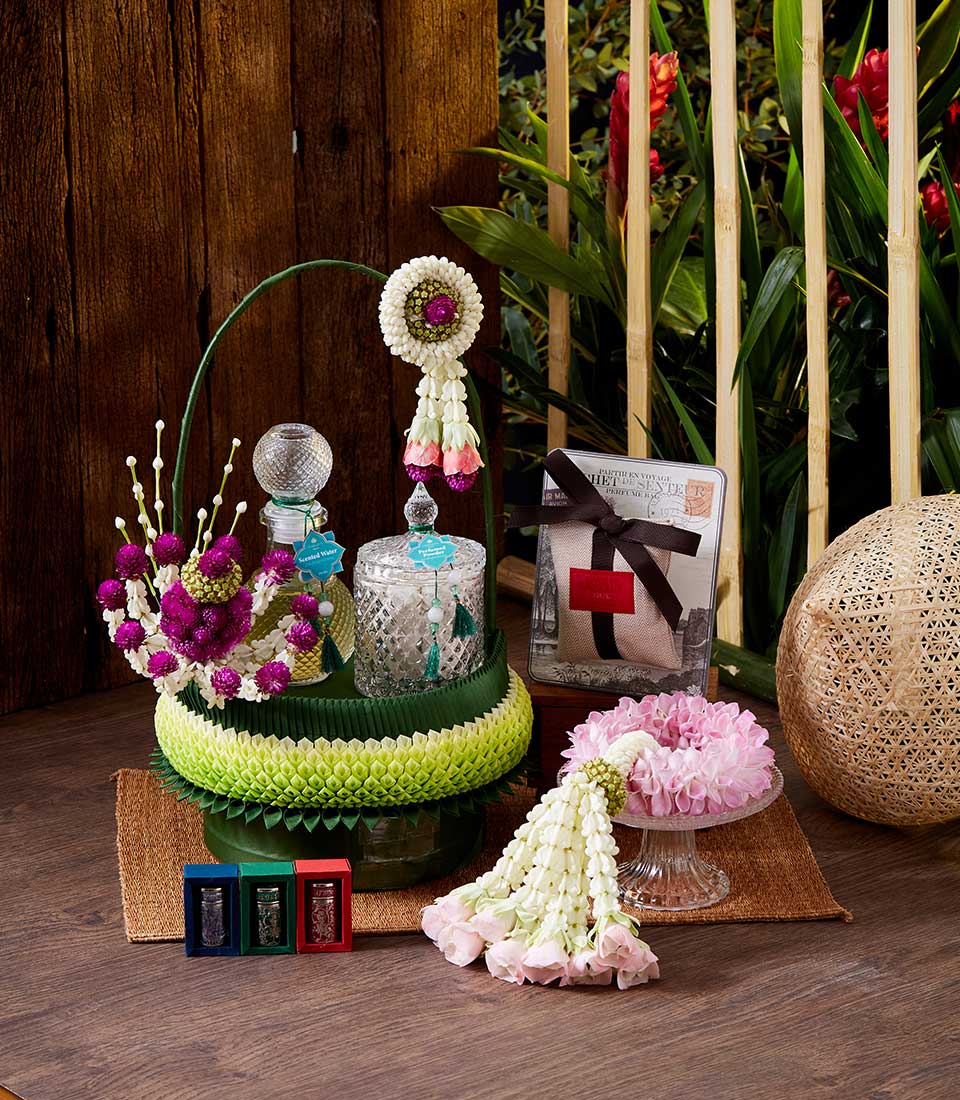A display of elegant floral arrangements and decorative items is placed on a wooden surface. The center features glass bottles with ornate flower decorations and a Songkran Thai incense set. Surrounding them is a floral garland, a pink flower bouquet, three small boxes, and a neatly wrapped gift.
