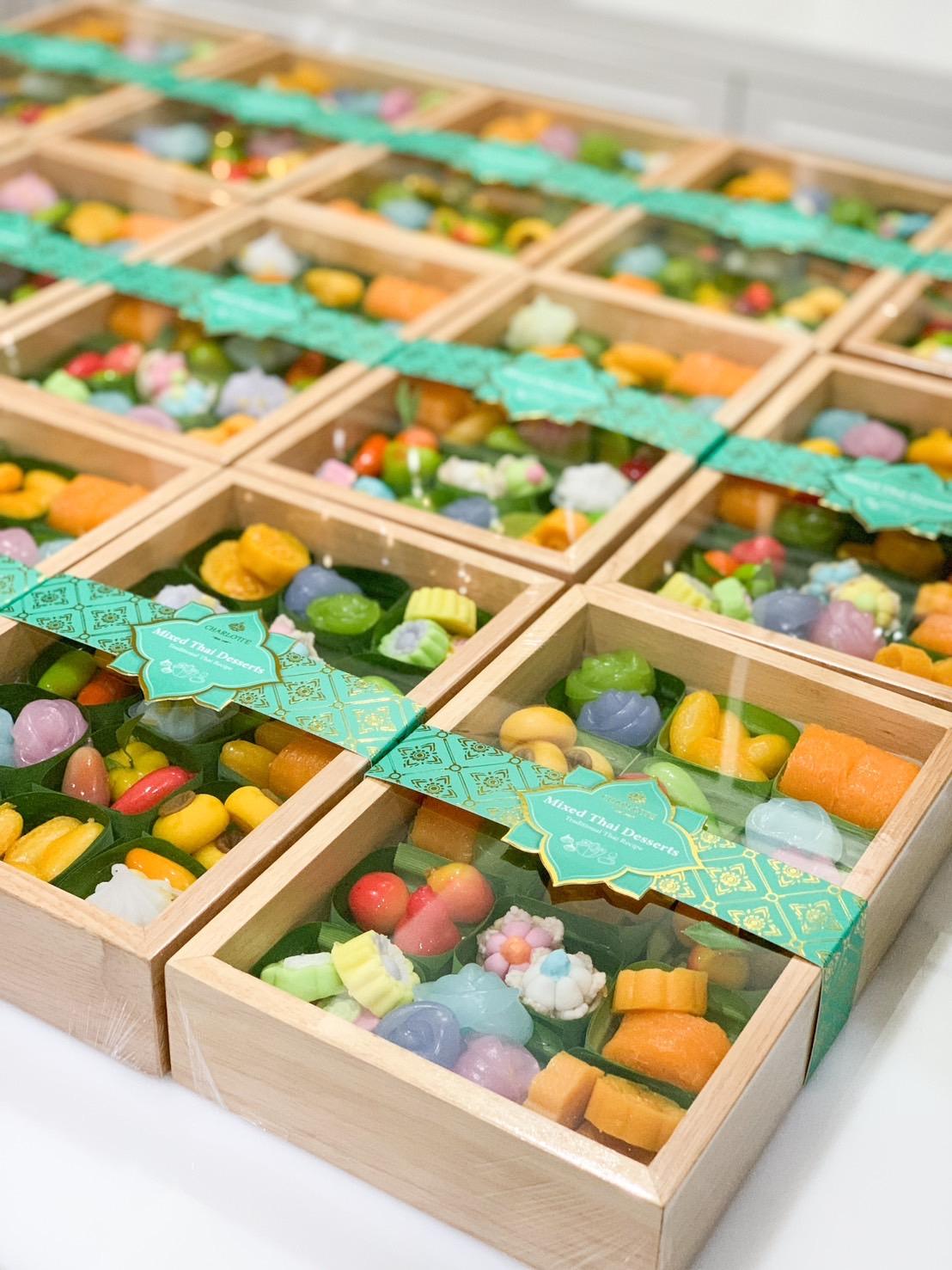 Rows of wooden boxes filled with colorful, assorted Thai sweets, each box wrapped with a green, patterned ribbon. The vibrant sweets come in various shapes and sizes, neatly arranged inside the transparent lids. This charming display could be part of a delightful Songkran set.