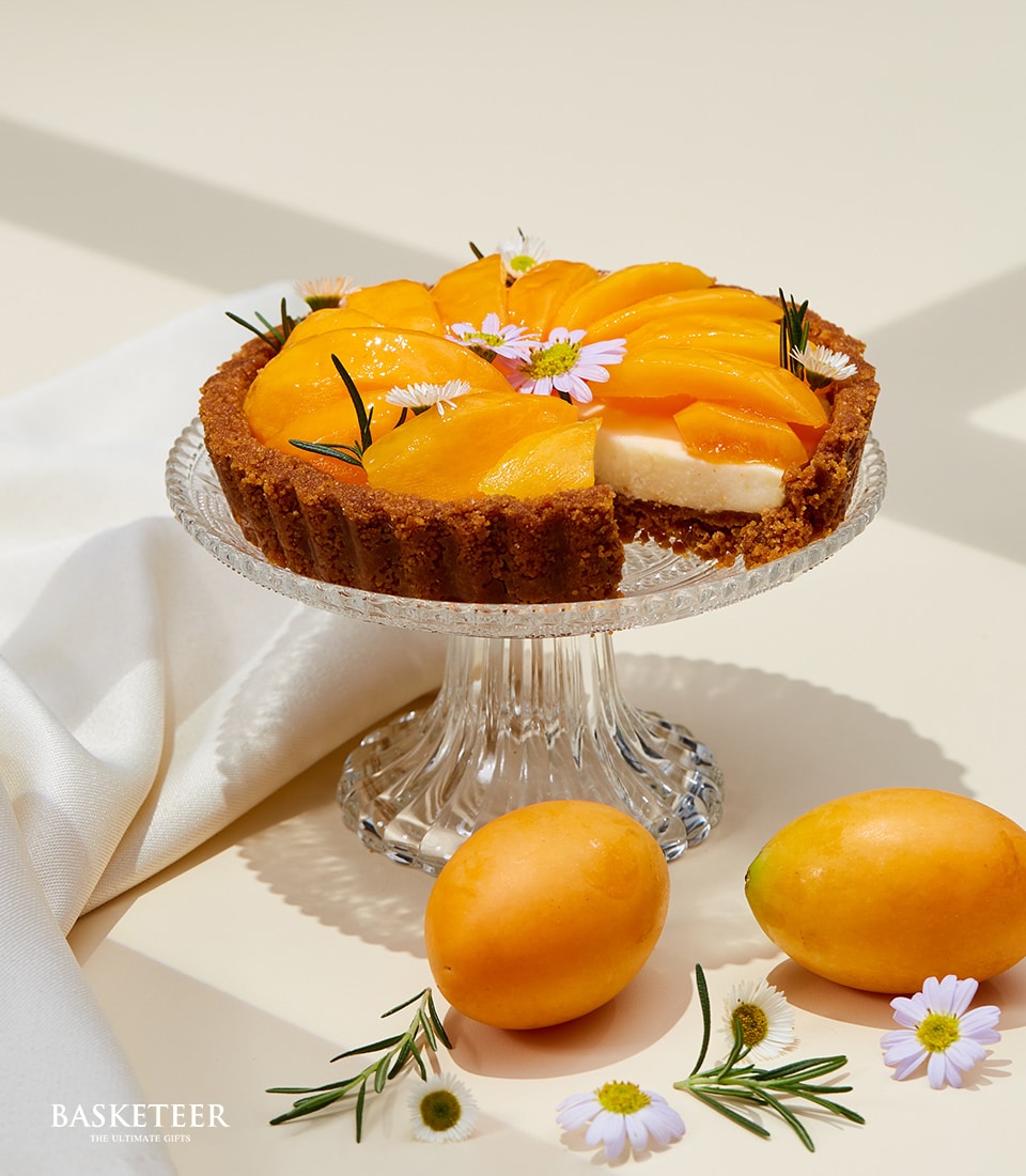 A mouthwatering Mayongchid Cheese Tart by Basketeer gracefully atop a gleaming glass pedestal, set against an elegantly understated background. The tart is adorned with delicately sliced peaches, petite blossoms, and aromatic rosemary sprigs. Surrounding the pedestal, you’ll find two perfectly ripe whole peaches and artfully scattered rosemary and flowers. A touch of sophistication is added with the striking Basketeer branding discreetly present in the scene.