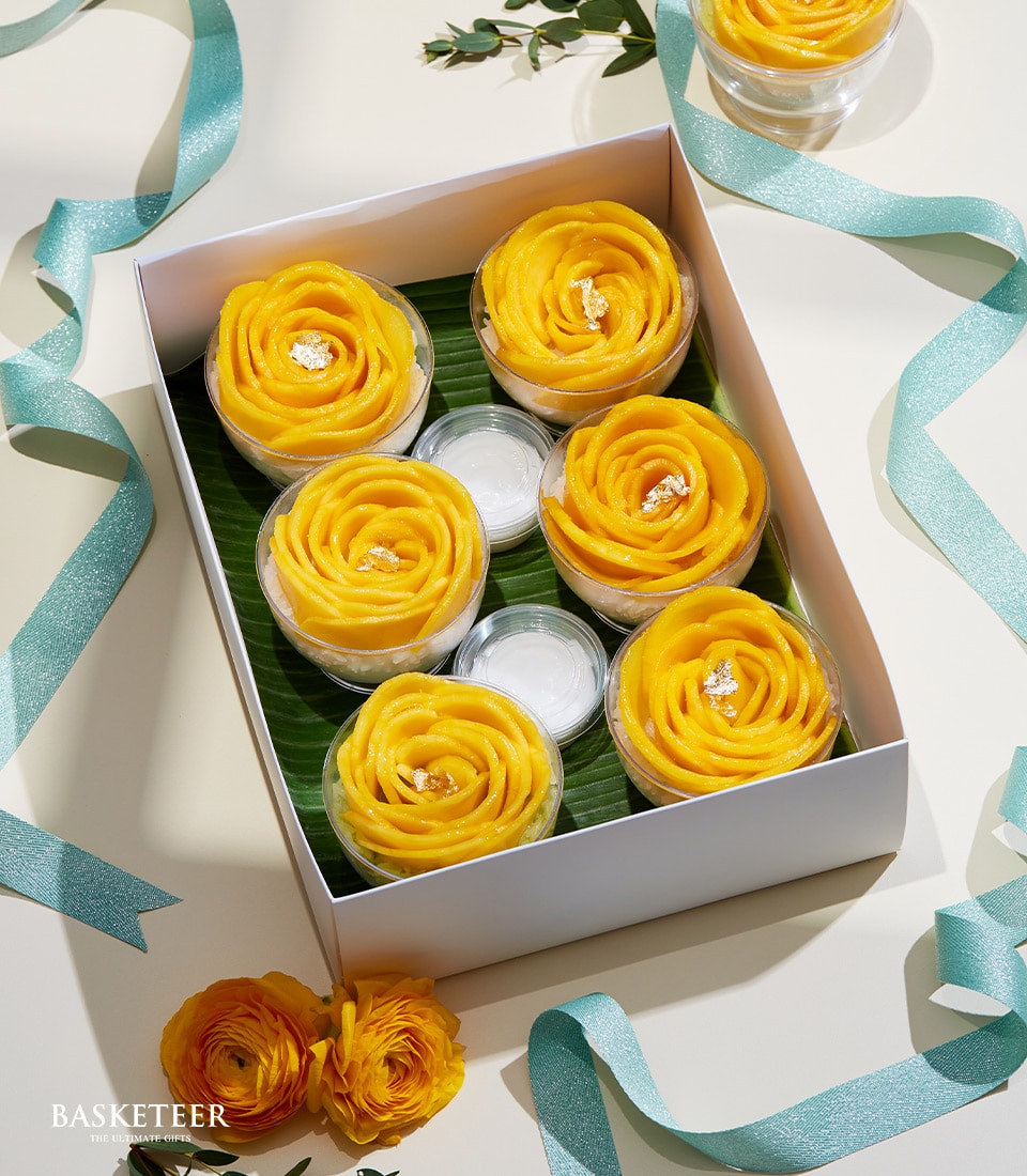 A luxurious white box showcases an exquisite arrangement of six rose-shaped desserts, meticulously crafted from golden mango slices, evoking the elegance of a premium Mango Sticky Rice Set. Accompanying the delicacies are two petite white containers, likely holding velvety sauce or cream for an enhanced tasting experience. Teal ribbons and vibrant yellow ranunculus flowers gracefully frame this sumptuous treat, while the prestigious 