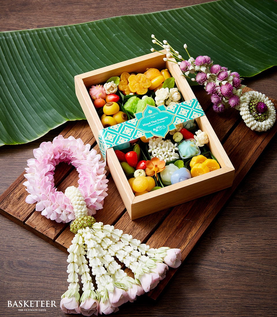 A sumptuous wooden box brimming with an array of vibrant Thai sweets graces a pristine wooden board. Surrounding this lavish assortment are delicately arranged flower garlands and an expansive, lush green banana leaf, perfectly capturing the essence of Songkran. The box is elegantly adorned with a ribbon featuring the 