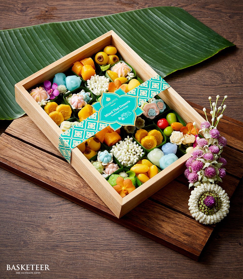 A luxurious wooden box from Basketeer, brimming with an exquisite array of colorful Thai sweet delicacies, rests elegantly on a polished wooden surface. The box is adorned with a sophisticated blue decorative band, proudly displaying the Basketeer label. Enhancing its allure, two pieces of fresh banana leaf lie gracefully beside the box, complemented by a handmade floral garland. This meticulously curated set makes for an impeccable Songkran gift that exudes elegance and thoughtful charm.