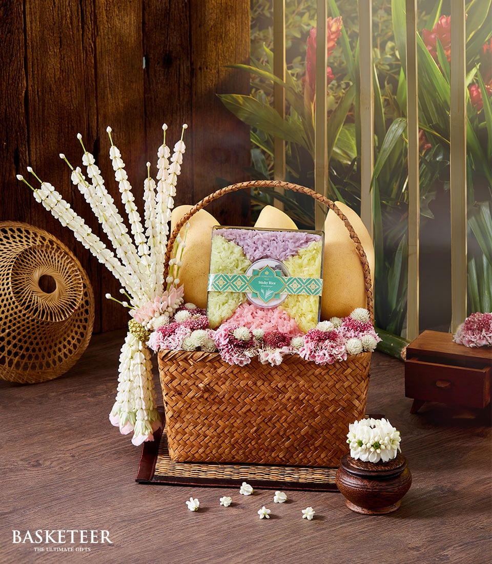 A luxurious woven basket, brimming with pastel-colored blossoms and plush beige cushions, graces a wooden surface against the backdrop of a verdant garden. The exquisite Thai Floral Mango Basket features an elegantly patterned box, accompanied by a delicate garland of pristine white flowers draped gracefully along its side. The prestigious 