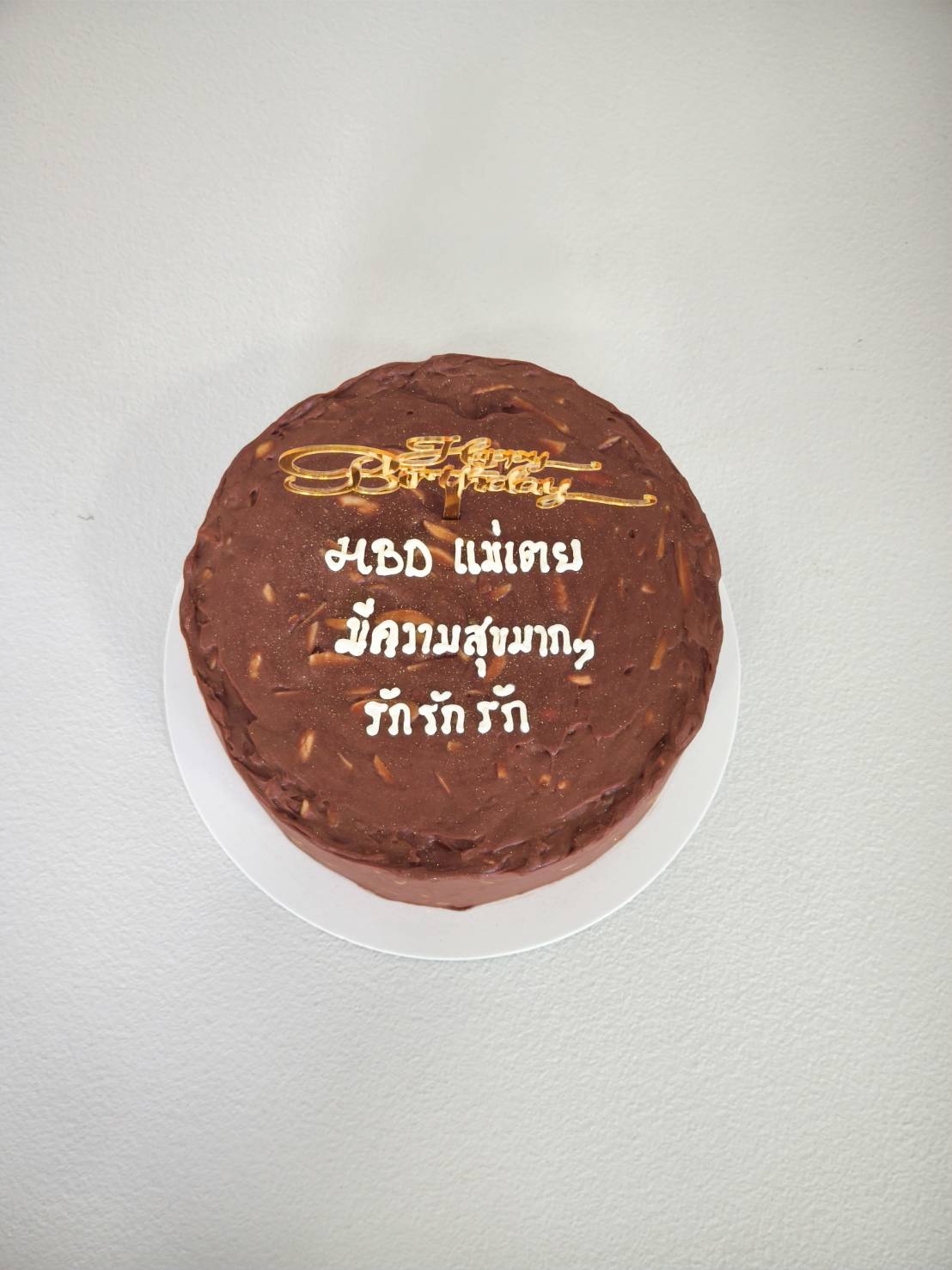 Round chocolate birthday cake with almond chocolate frosting, gold “Happy Birthday” topper, and personalised Thai message written in white icing.
