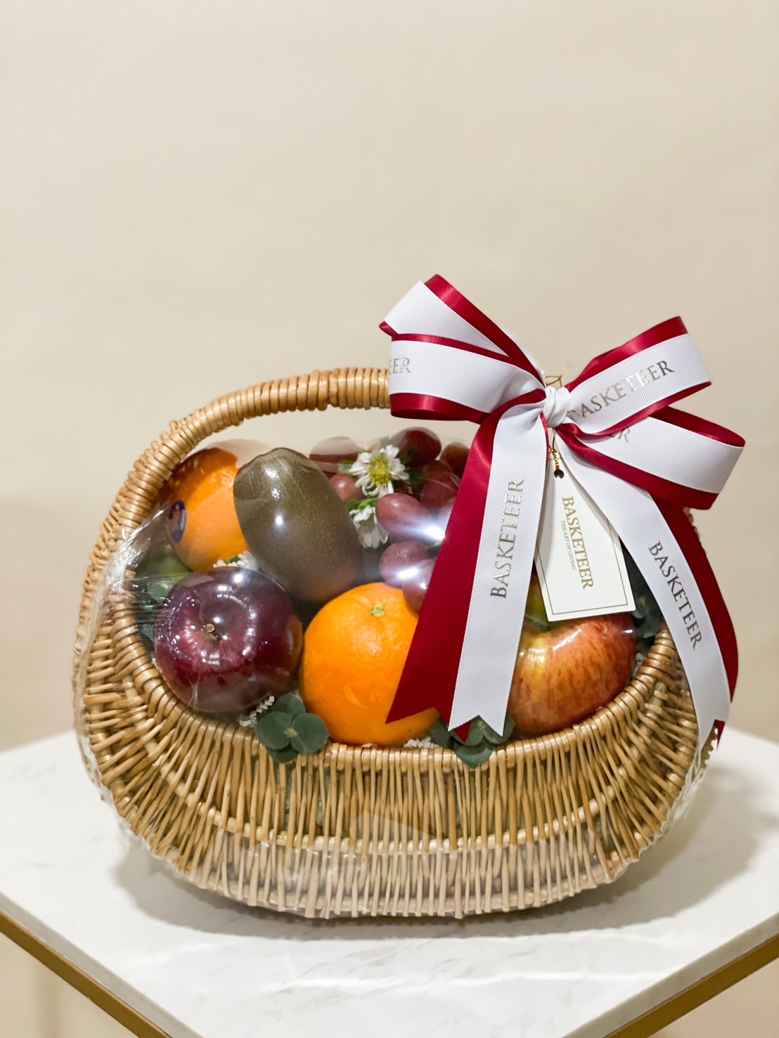 Wicker basket filled with fresh fruits like apples, oranges, and grapes, adorned with a red and white ribbon by Basketeer.