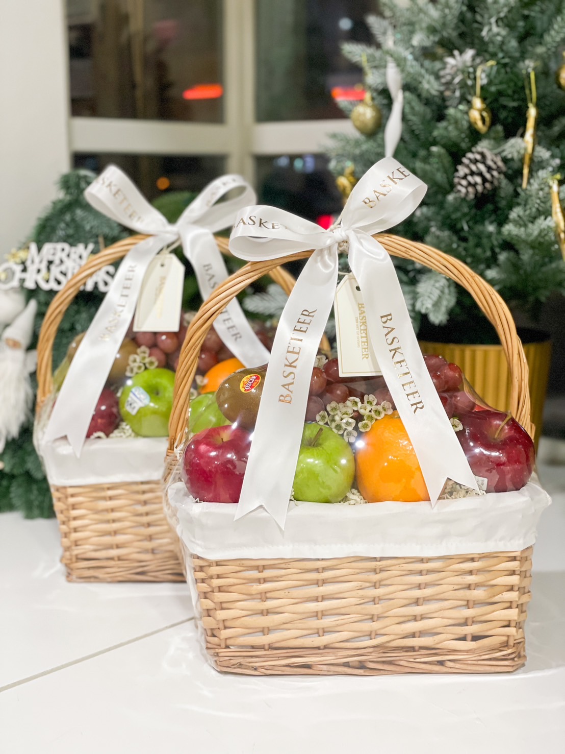 Set of wicker fruit baskets filled with apples, oranges, grapes, and pears, elegantly tied with white ribbons by Basketeer.