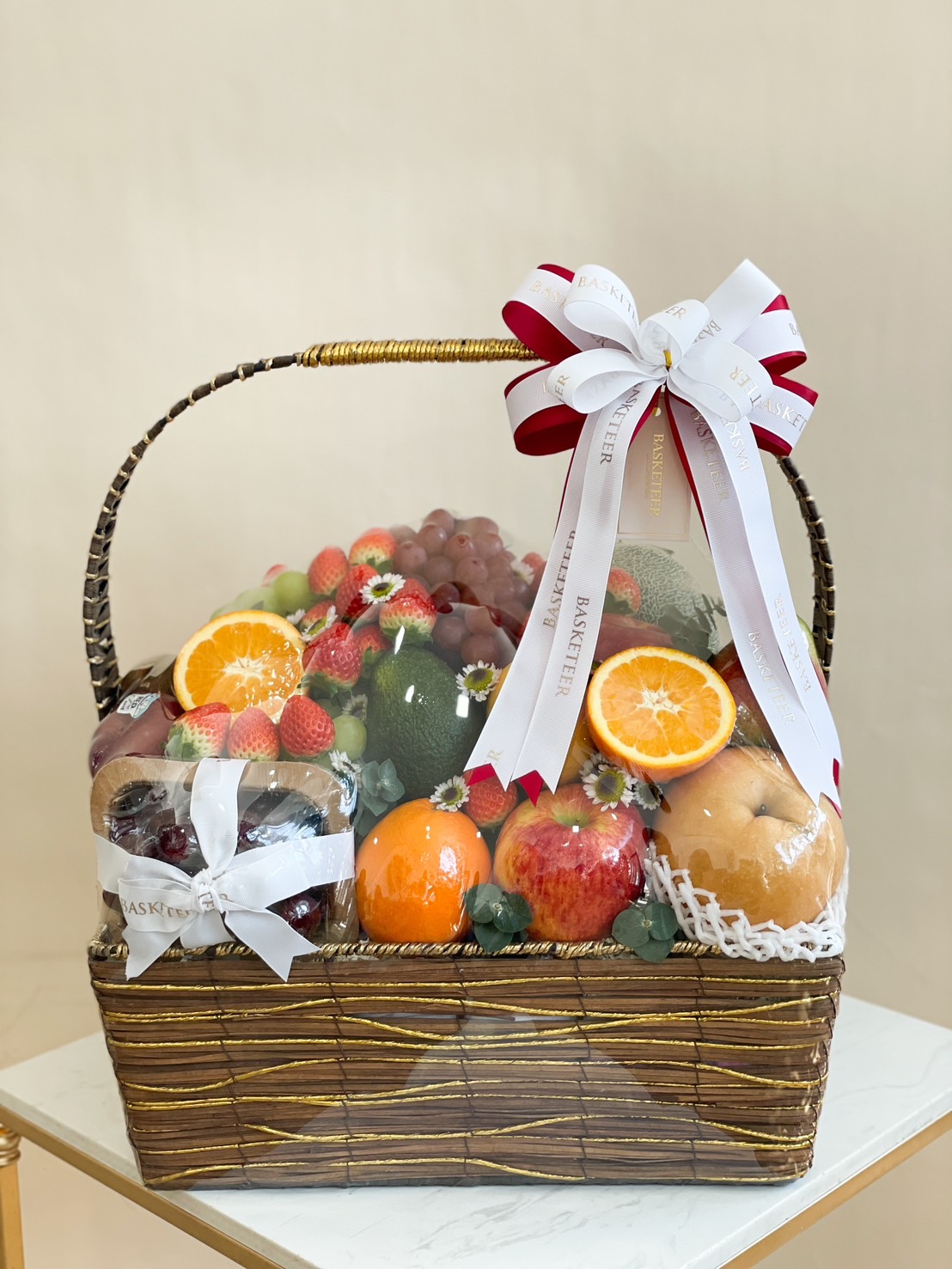 A wicker fruit basket containing fresh apples, oranges, strawberries, grapes, cherries, and avocados, tied with a white and red decorative ribbon.