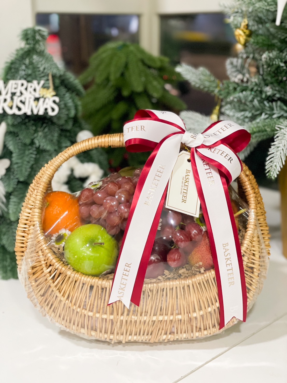A beautifully crafted wicker basket filled with apples, oranges, grapes, and strawberries, adorned with a red and white Basketeer ribbon.