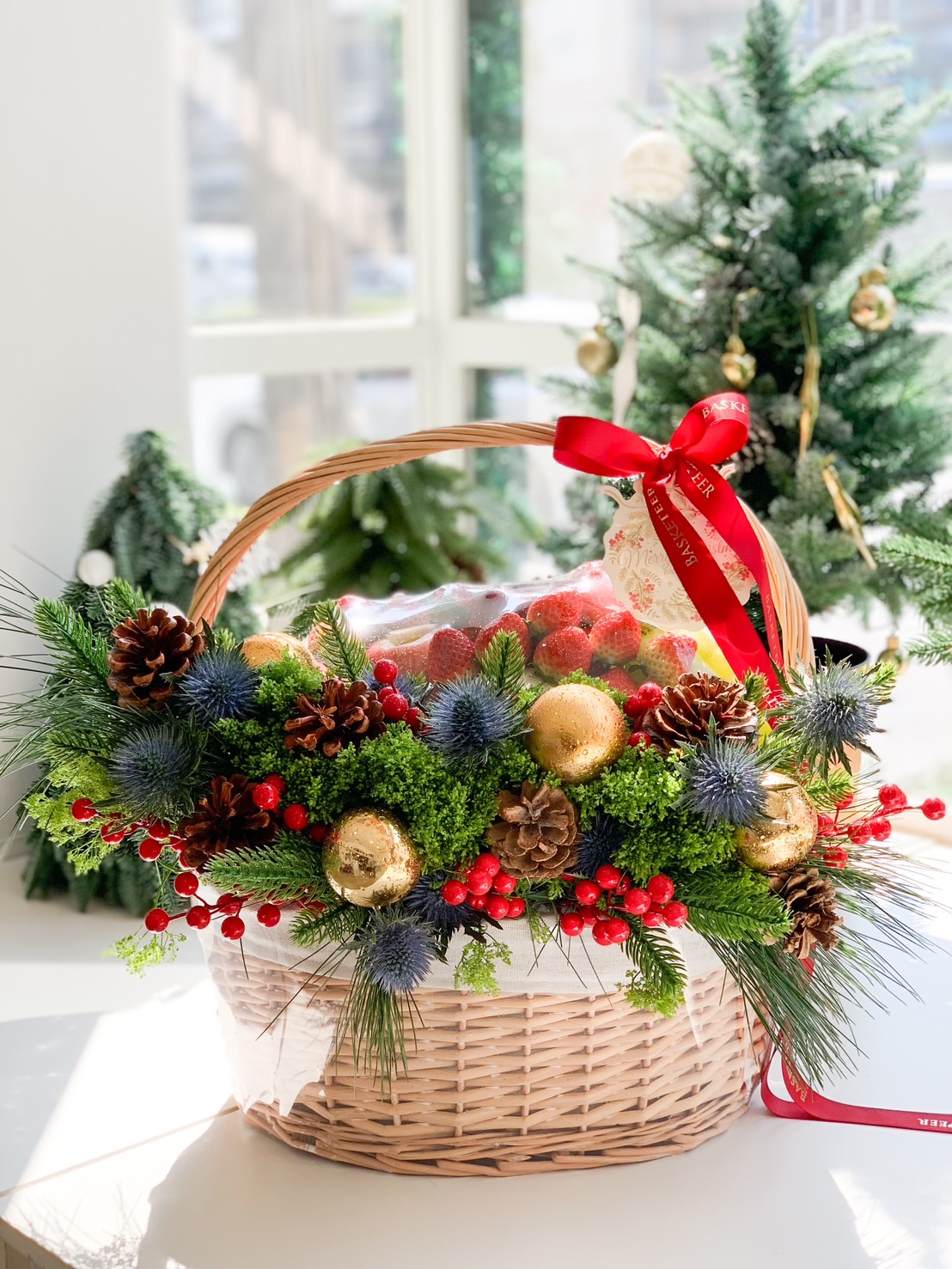 Festive holiday fruit basket featuring fresh strawberries, pinecones, vibrant greenery, red berries, and golden ornaments, tied with a red ribbon.