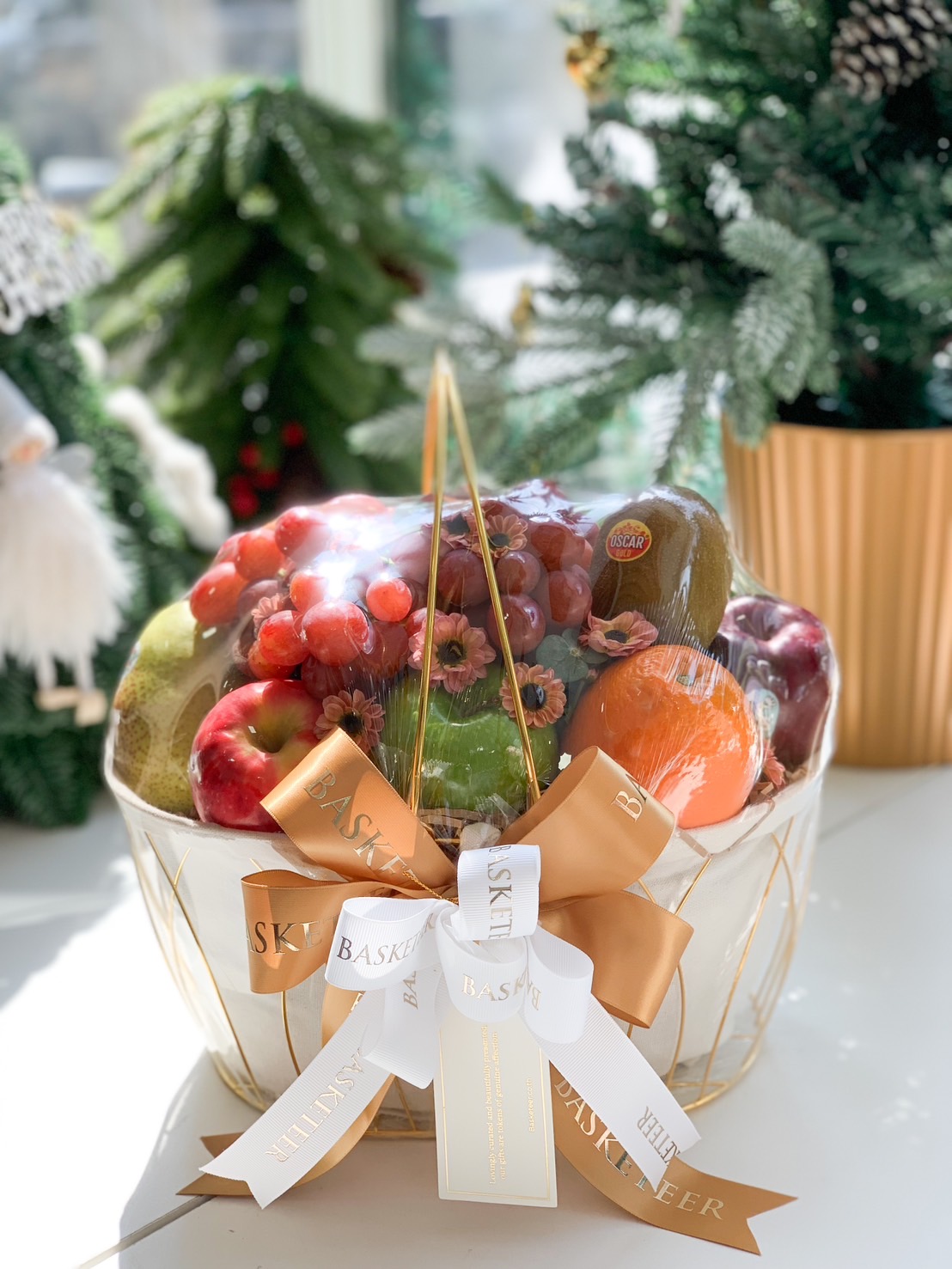 Premium fresh fruit basket containing apples, oranges, kiwis, and grapes, beautifully wrapped with a golden and white ribbon, displayed near a festive Christmas tree.