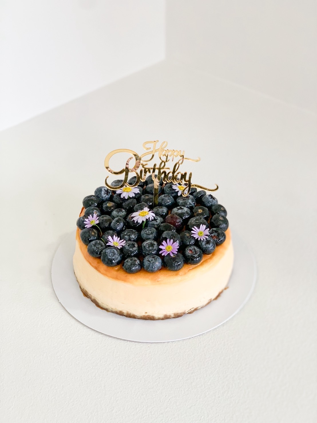 Fresh blueberry cheesecake topped with blueberries and mini purple flowers with Happy Birthday topper, premium birthday cake.
