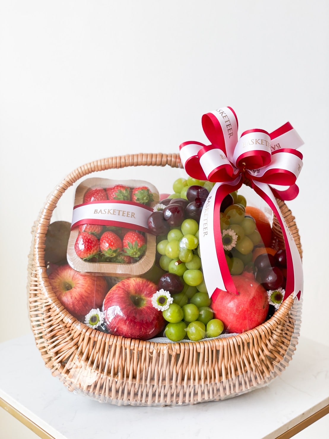 A large wicker basket filled with a mixed fruit selection, including red and green grapes, strawberries in a plastic container, and apples. The basket is adorned with a white and red ribbon labeled 